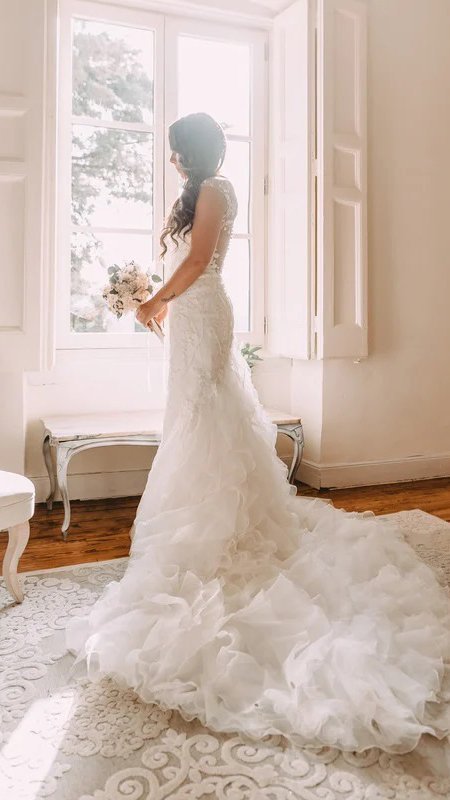 A bride in a wedding dress standing gracefully in front of a window.