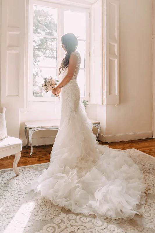 A bride in a wedding dress standing in front of a window. Wedding gown by John Emily Studio.