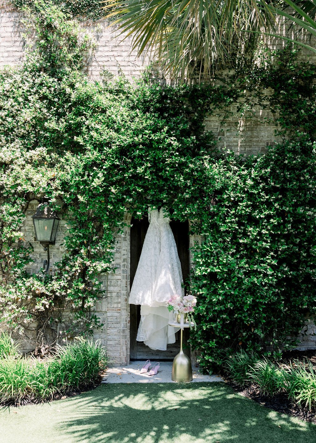 A lace wedding gown hanging in an ivy-covered doorway with pink shoes below