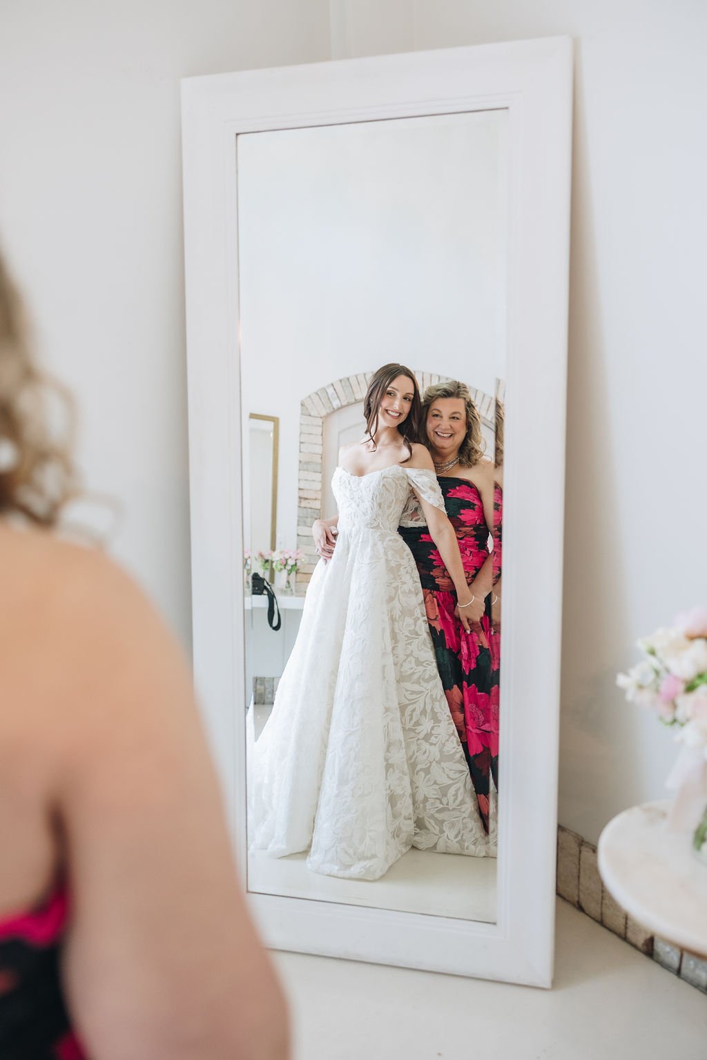 Bride and a woman in a pink floral formal dress reflected in a mirror